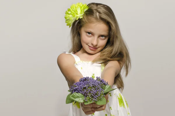 Beautiful young blonde girl in white dress smiling, portrait of an adorable kid. Little girl with a green big flower in her hair, smelling a flower — Stock Photo, Image