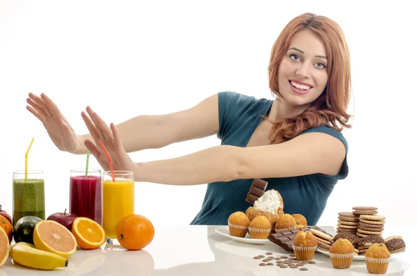 Женщина выбирает между фруктами, смузи и органической здоровой пищи против сладостей, сахара, много конфет, нездоровой пищи. Лечение кондитерской зависимости с фруктами и овощами — стоковое фото