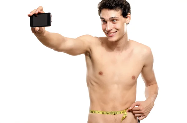 Skinny man taking a selfie with his phone while measuring his waist with a centimeter. Beautiful teenager taking a photo for facebook.Anorexic young man training to become stronger — Stock Photo, Image