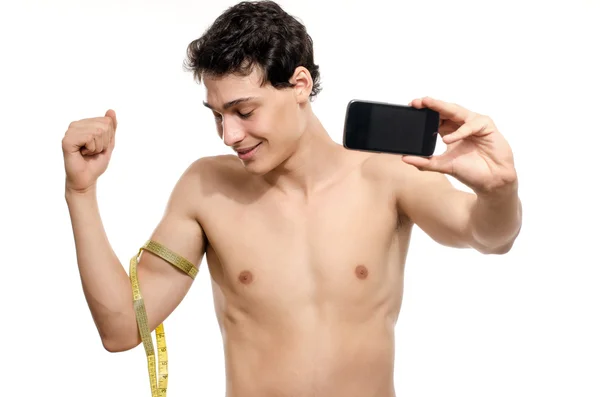 Skinny man taking a selfie with his phone while measuring his biceps with a centimeter. Beautiful teenager taking a photo for facebook.Anorexic young man training to become stronger — Stock Photo, Image