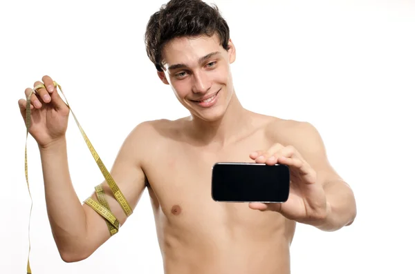 Skinny man taking a selfie with his phone while measuring his biceps with a centimeter. Beautiful teenager taking a photo for facebook.Anorexic young man training to become stronger — Stock Photo, Image