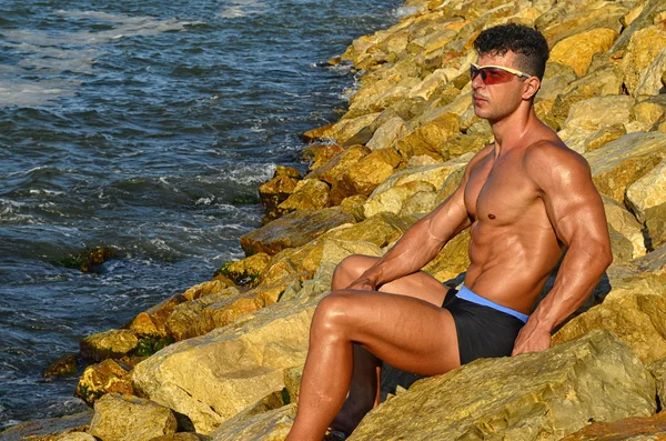 Strong bodybuilder with six pack.Fitness trainer with perfect abs, shoulders,biceps, triceps,chest, flexing his muscles on the beach with sea waves on the background, training in vacation