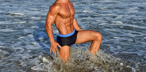 Forte bodybuilder con sei pacchetti.Fitness trainer con addominali perfetti, spalle, bicipiti, tricipiti, petto, flettendo i muscoli sulla spiaggia con onde marine sullo sfondo, allenamento in vacanza — Foto Stock