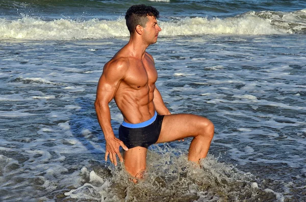 Forte bodybuilder con sei pacchetti.Fitness trainer con addominali perfetti, spalle, bicipiti, tricipiti, petto, flettendo i muscoli sulla spiaggia con onde marine sullo sfondo, allenamento in vacanza — Foto Stock