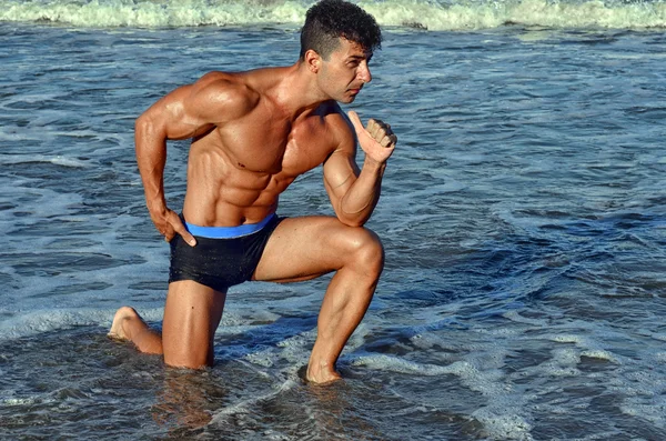 Culturista fuerte con seis pack.Fitness entrenador con abdominales perfectos, hombros, bíceps, tríceps, pecho, flexionar sus músculos en la playa con olas de mar en el fondo, entrenamiento en vacaciones —  Fotos de Stock