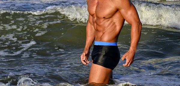 Starker Bodybuilder mit sechs Packungen. Fitness-Trainer mit perfektem Bauch, Schultern, Bizeps, Trizeps, Brust, lässt seine Muskeln am Strand mit Meereswellen im Hintergrund spielen, Training im Urlaub — Stockfoto