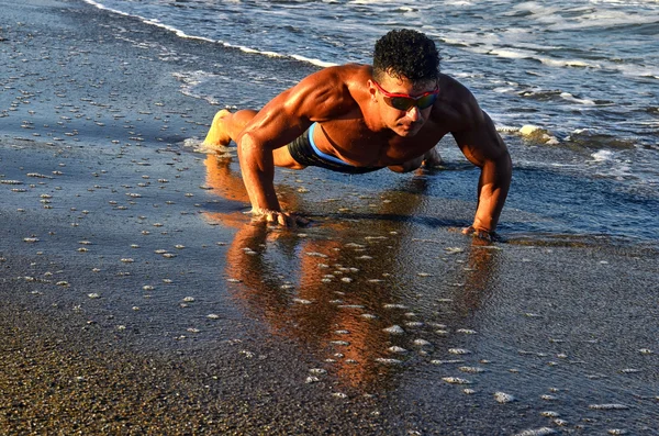 Strong bodybuilder with six pack.Fitness trainer with perfect abs, shoulders,biceps, triceps,chest, flexing his muscles on the beach, training in vacation, doing push ups in the sand — Zdjęcie stockowe