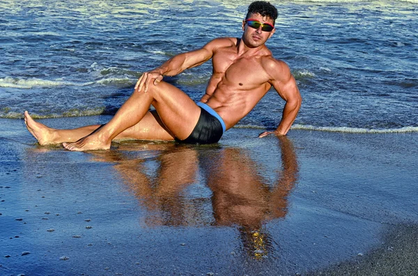 Forte bodybuilder con sei pacchetti.Fitness trainer con addominali perfetti, spalle, bicipiti, tricipiti, petto, flettendo i muscoli sulla spiaggia con onde marine sullo sfondo, allenamento in vacanza — Foto Stock