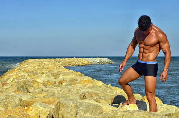 Forte bodybuilder con sei pacchetti.Fitness trainer con addominali perfetti, spalle, bicipiti, tricipiti, petto, flettendo i muscoli sulla spiaggia con onde marine sullo sfondo, allenamento in vacanza — Foto Stock