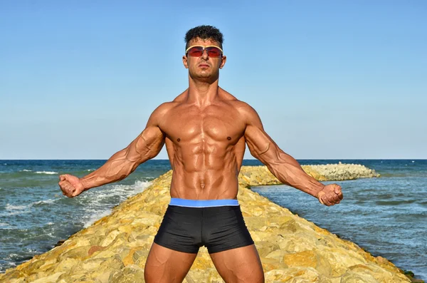 Strong bodybuilder with six pack.Fitness trainer with perfect abs, shoulders,biceps, triceps,chest, flexing his muscles on the beach with sea waves on the background, training in vacation — ストック写真
