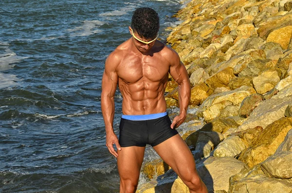 Strong bodybuilder with six pack.Fitness trainer with perfect abs, shoulders,biceps, triceps,chest, flexing his muscles on the beach with sea waves on the background, training in vacation — Stock Photo, Image