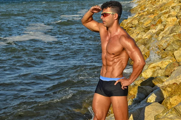 Culturista fuerte con seis pack.Fitness entrenador con abdominales perfectos, hombros, bíceps, tríceps, pecho, flexionar sus músculos en la playa con olas de mar en el fondo, entrenamiento en vacaciones —  Fotos de Stock