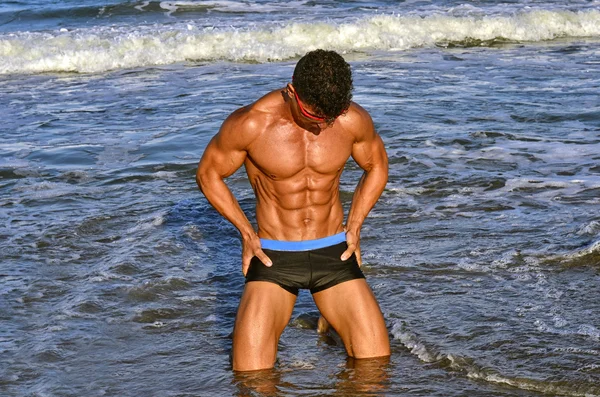 Starker Bodybuilder mit sechs Packungen. Fitness-Trainer mit perfektem Bauch, Schultern, Bizeps, Trizeps, Brust, lässt seine Muskeln am Strand mit Meereswellen im Hintergrund spielen, Training im Urlaub — Stockfoto