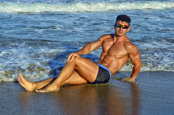 Strong bodybuilder with six pack.Fitness trainer with perfect abs, shoulders,biceps, triceps,chest, flexing his muscles on the beach with sea waves on the background, training in vacation — Stock Photo, Image