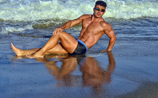 Strong bodybuilder with six pack.Fitness trainer with perfect abs, shoulders,biceps, triceps,chest, flexing his muscles on the beach with sea waves on the background, training in vacation — Stock Photo, Image