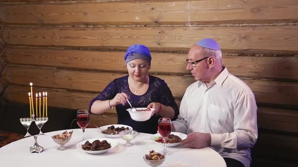 Zsidó házaspár egy feleség fejdísszel egy férfi egy kippah-ban egy asztalnál gyertyákkal a Hanuka vacsorák napján.. — Stock videók