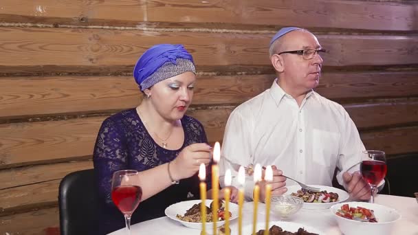 Еврейская супружеская пара жена в головном уборе мужчина в киппе за столом с горящими свечами во дни Хануки обедает — стоковое видео