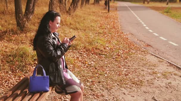 Eine schwangere Frau in einer Gasse auf einer Bank arbeitet im Herbst mit einem Smartphone — Stockvideo