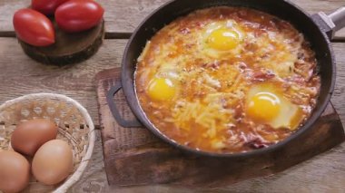 Bir el, ahşap bir standın üstündeki masaya bir tava ve Shakshuka koyar.