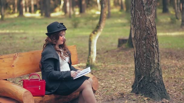 Uma mulher com cabelo castanho em um chapéu em botas vermelhas e uma bolsa vermelha em um banco de parque no outono lê — Vídeo de Stock