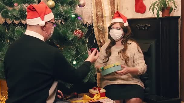 Egy családi férj és feleség karácsony reggelén, álarcot viselve járvány idején, ajándékokat fontolgatnak a karácsonyfán. — Stock videók