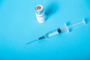Mavi arka planda, bir şırınga ve bir şişe koronavirüs aşısı..