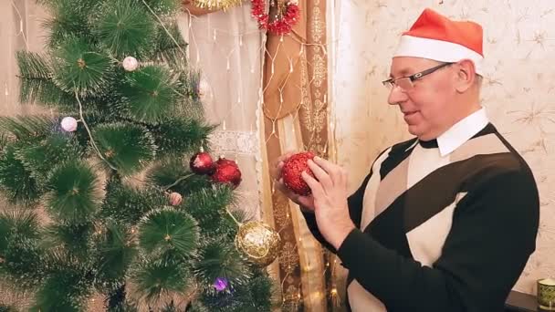 Um homem de chapéu de Pai Natal decora uma árvore de Natal com bolas — Vídeo de Stock