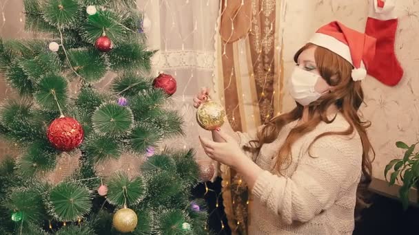 Una mujer con una máscara médica y un sombrero de Santa Claus se prepara para la Navidad durante una epidemia y decora un árbol de Navidad con juguetes — Vídeos de Stock