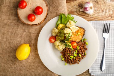 Brokolili fırınlanmış balık ve biberiye ve limonlu beyaz bir tabakta mercimek bir çatal ve sebzenin yanında kareli peçete. Yatay fotoğraf