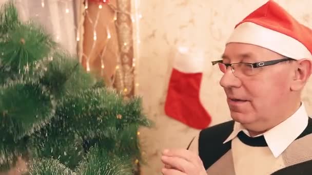 Un hombre con un sombrero de Santa Claus recoge un árbol de Navidad artificial en casa — Vídeos de Stock