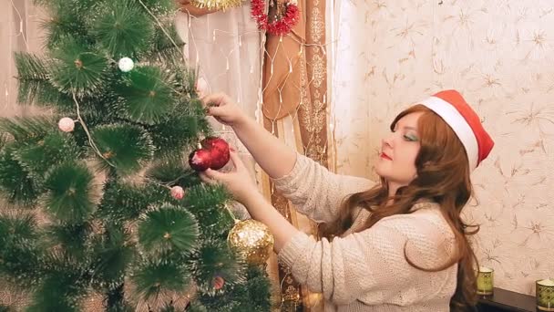 Een jonge vrouw in een kerstmuts versiert een kerstboom met ballen — Stockvideo