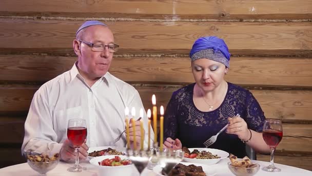 Una pareja judía casada en tocados tradicionales come y bebe vino en una mesa festiva con velas encendidas durante Hanukkah — Vídeo de stock