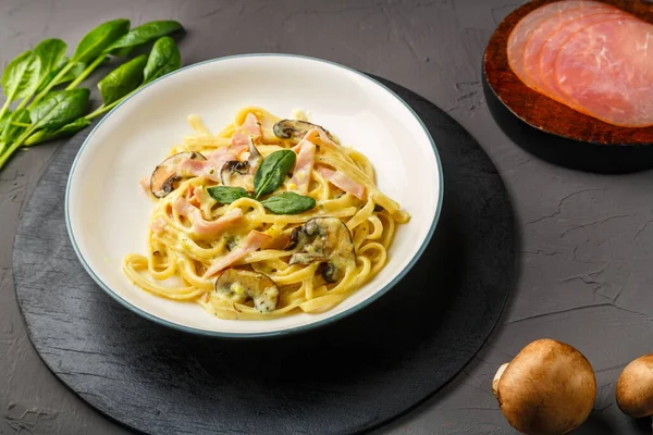 Kremalı sosta jambon ve mantarlı bir tabak makarna siyah yuvarlak bir tezgahta fesleğenle birlikte mantar ve jambonun yanında çatal. Yatay fotoğraf.