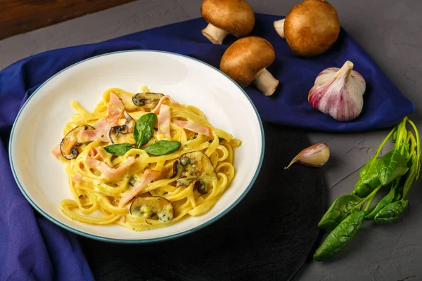 Kremalı sosta jambon ve mantarlı bir tabak makarna ve siyah yuvarlak bir stantta fesleğenle sarımsak ve otların yanında mavi bir peçetede çatal. Yatay fotoğraf.