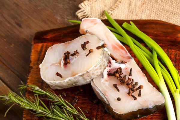 Steaks de morue blanche fraîche sur une planche de bois saupoudrée d'épices à côté de romarin et d'oignons. — Photo