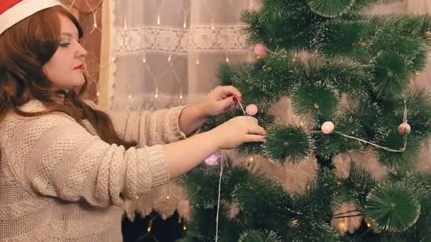 Žena v klobouku Santa Claus maluje vánoční stromek s elektrickými lampami. — Stock video