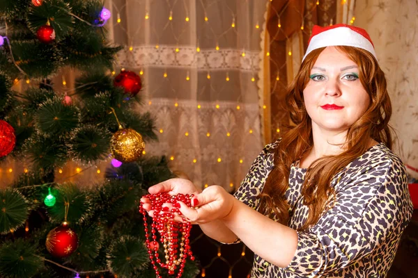Una mujer pelirroja en un sombrero Santau árbol de Navidad sostiene decoraciones de cadena en sus manos. — Foto de Stock