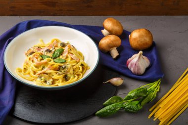 Kremalı sosta jambon ve mantarlı bir tabak makarna. Siyah bir tezgahta fesleğen ve sarımsak ve otların yanında mavi bir peçetede çatal..