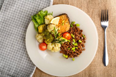 Brokolili fırında balık ve biberiye ve limonlu beyaz bir tabakta mercimek peçetenin üstünde ve çatalın yanında yuvarlak bir tezgah..