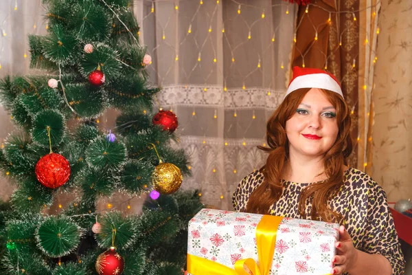 Una mujer en un sombrero de Santa Claus cerca de un árbol decorado de Navidad sostiene una caja grande con un regalo. — Foto de Stock