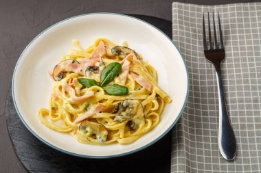 Kremalı sosta jambonlu ve mantarlı bir tabak makarna siyah yuvarlak bir tezgahta fesleğenle peçetede bir çatalla.