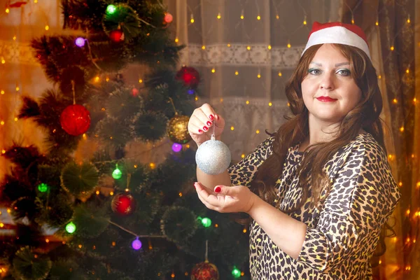 Una mujer de cabello castaño en un sombrero de Santa junto a un árbol de Navidad con una bola de decoración en las manos. — Foto de Stock