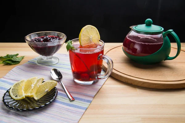 Berry tea in a cup next to a teapot spoon and lemon and lemon balm and jam in a vase on a round stand and a napkin. — Stock Photo, Image