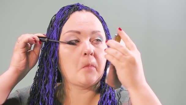 Eine Frau mit blauer Afro-Frisur bemalt ihre Augen mit Wimperntusche und blickt zu Beginn des Arbeitstages in einen kleinen Spiegel — Stockvideo