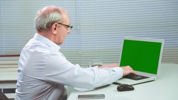 Um homem com óculos em um escritório com persianas em um computador imprime uma tela verde e escreve em um caderno — Vídeo de Stock