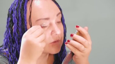 Ofisteki mavi afro saçlı bir kadın küçük bir aynaya bakan gölgeli gözleri boyuyor. Ortam planı