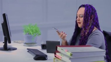 Ofiste mavi afro örgülü bir kadın bilgisayar başında telefonla iletişim kuruyor. Genel plan