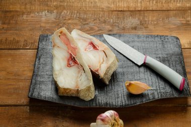 Siyah ahşap bir tahtanın üzerinde taze balık bifteği bıçak ve sarımsağın yanında. Yatay fotoğraf