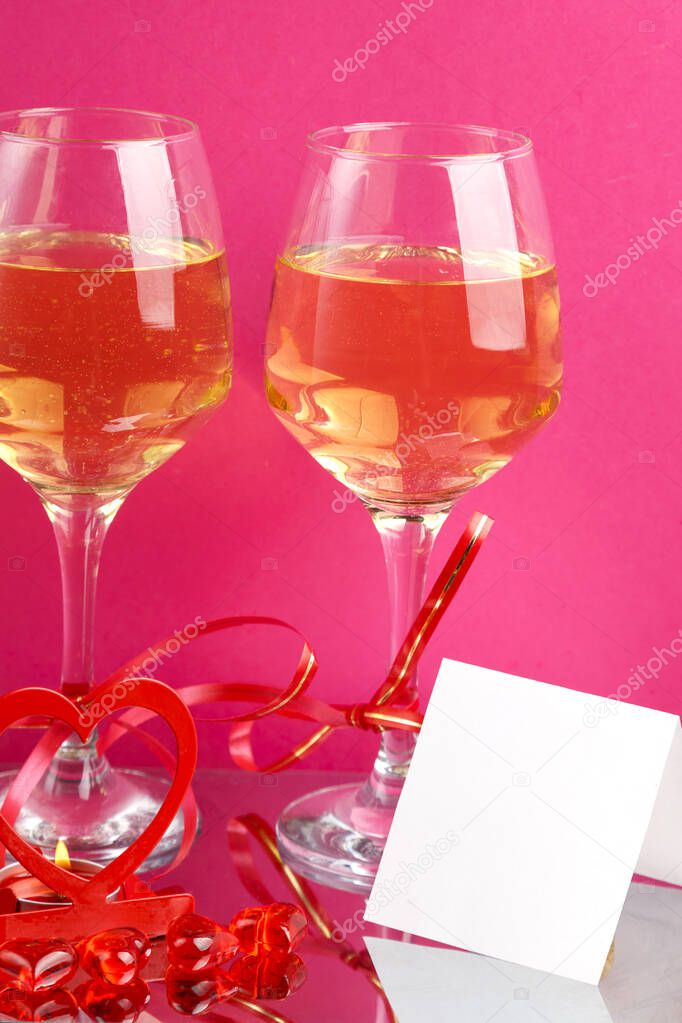 Two glasses with champagne tied with red ribbons on a pink background next to a postcard. Vertical photo