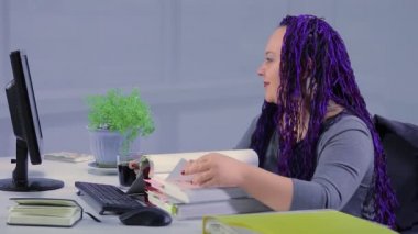 Ofiste mavi afro örgülü bir kadın raporlara bakıyor ve video bağlantısıyla iletişim kuruyor. Genel plan